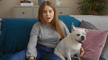 Genç, güzel, sarışın bir kadın, beyaz köpeğiyle oturma odasında oturuyor. Yüzünde şaşırmış bir ifade var..