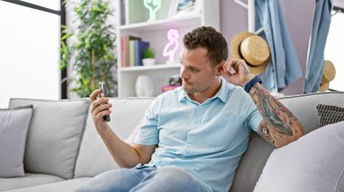 Evdeki gri koltukta oturan yakışıklı adam telefonuna tuhaf bir ifadeyle bakıyor..