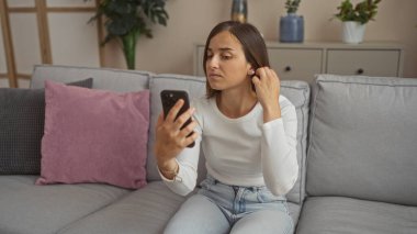 Oturma odasındaki bir kanepede oturan genç bir kadın, odaklanmış bir ifadeyle küpesini düzeltirken telefonuna bakıyor, rahat bir yastık ve kapalı alan bitkilerine karşı kurulmuş..