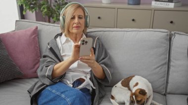 Akıllı telefon kullanan olgun bir sarışın kadın kulaklık takıyor ve modern bir oturma odasında köpeğiyle koltukta oturuyor..