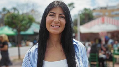 Güneşli bir günde arka planda insanlarla birlikte canlı bir şehir ortamında gülümseyen güzel, genç, İspanyol bir esmer kadın..