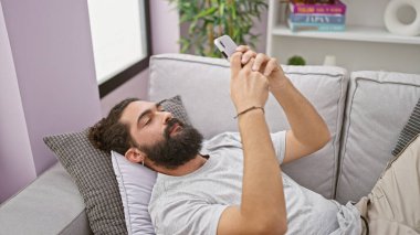 Sakallı İspanyol bir adam rahat bir oturma odasında kanepede dinlenirken akıllı telefon kullanıyor..