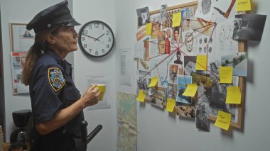 Elinde kahve fincanı olan orta yaşlı bir kadın polis memuru ofis benzeri bir odada kanıt duvarını inceliyor..