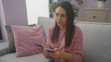 Genç bir İspanyol kadın kredi kartını evin içinde tutuyor, rahat ve iyi dekore edilmiş bir dairede bir koltukta oturuyor..