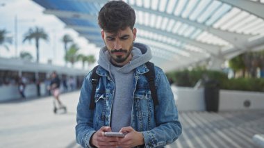 Sakallı genç bir İspanyol, şehir caddesinde dikilirken akıllı telefon kullanıyor..