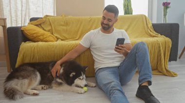İspanyol adam, rahat oturma odasında Husky 'yi okşarken akıllı telefon kullanıyor.