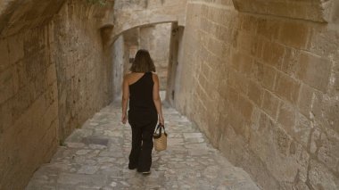 Genç bir İspanyol kadın, Mata, Basilicata, İtalya 'nın büyüleyici eski kasabasında yürür, her iki tarafında antik duvarlar olan dar, taştan bir sokak boyunca bir sepet tutar..