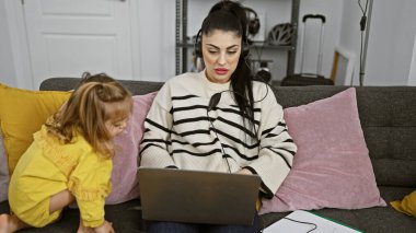 Kulaklıklı, oyuncakçı kızıyla birlikte rahat bir oturma odasında dizüstü bilgisayar kullanan odaklanmış bir kadın.