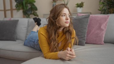 Genç, güzel, sarışın bir kadın, yumuşak yastıklar ve yeşil bitkilerle çevrili, modern bir oturma odasında rahat bir kanepeye uzanıyor..