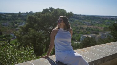 Beyaz elbiseli güzel bir İspanyol kadın güneşli bir günün tadını çıkarıyor. Taş bir duvarda oturuyor. Locorotondo manzaralı, puglia 'nın yemyeşil manzaralı..
