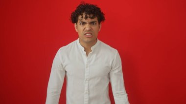 Beyaz tişörtlü hayal kırıklığına uğramış İspanyol bir adam canlı kırmızı bir arka planda izole edilmiş kafa karışıklığını ifade ediyor.