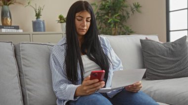 Genç esmer bir kadın oturma odasındaki bir kanepede oturuyor, bir elinde telefon, diğer elinde bir belge, odaklanmış ve düşünceli görünüyor..