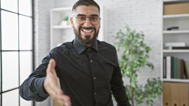 Profesyonel ortamda gülümseyen sakallı bir adam elini uzatıyor..