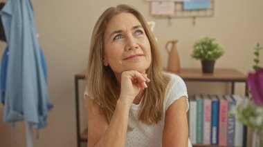 Düşünceli olgun bir kadın, arka planda ışık dekoru ve bitkilerle, sakin bir atmosferi çağrıştırarak, evde düşünmeye başlar..