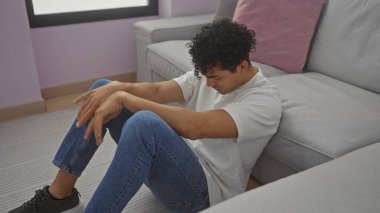 Gündelik giyinmiş İspanyol bir adam üzgün ya da düşünceli, modern gri bir kanepede oturuyor..