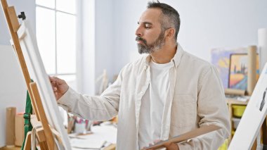 Normal kıyafetli olgun bir sakallı adam sanatsal malzemelerle çevrili bir sanat stüdyosundaki tuvale dikkatlice resim yapar..