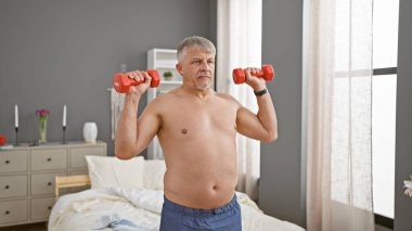 Olgun bir adam yatak odasında dambıllarla egzersiz yapıyor, evde sağlık ve spor yapıyor..