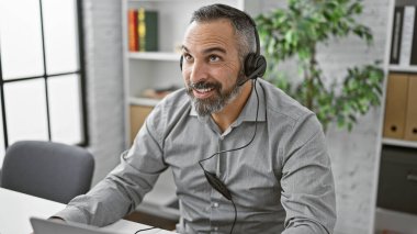 Modern ofiste bilgisayar başında çalışan, gülümseyen, kulaklıklı, yaşlı, İspanyol bir adam..