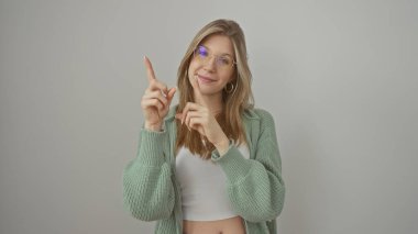 Gözlüklü genç sarışın bir kadın beyaz bir arkaplanı işaret ediyor.