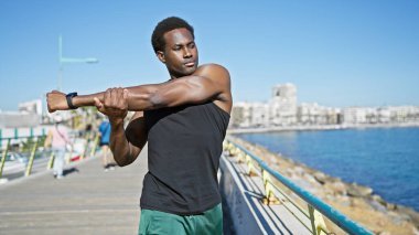 Açık mavi bir okyanus zeminiyle güneşli bir sahil geçidinde gerinen Afrikalı Amerikalı bir adam..