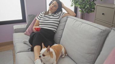 Bir kadın kanepede kulaklıklar, kahve fincanı ve sıcak bir oturma odasında bir chihuahua ile rahatlar..