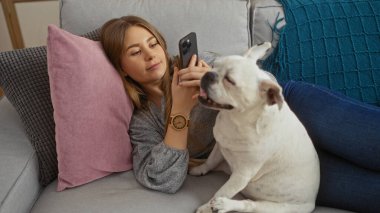 Genç sarışın bir kadın beyaz köpeğiyle kanepede rahat bir oturma odasında uzanırken akıllı telefonunu kullanıyor..