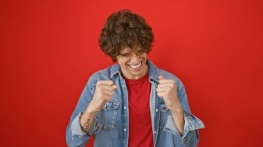 Sakallı, kırmızı arka planda kot giyen mutlu, genç İspanyol bir adam. Sıradan cazibe ve pozitiflik yayıyor..