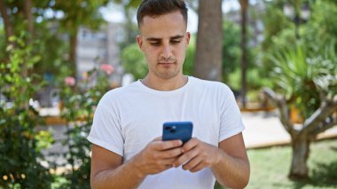 Yakışıklı genç bir adam yeşil bir parktaki akıllı telefonuna beyaz bir tişört giyerek mesaj atıyor..