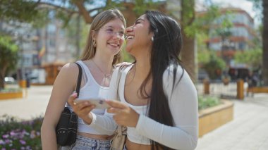 Ağaçlar ve binalarla çevrili güneşli bir şehir sokağında iki kadın gülüyor ve bir akıllı telefonu paylaşıyor.