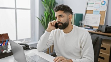 Modern ofis ortamında dizüstü bilgisayarla çalışan yakışıklı, sakallı adam, dalgın görünüyor..