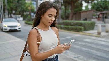 Zeki telefonuna cidden odaklanmış havalı İspanyol genç kadın neşeli bir şehrin göbeğinde güneşli sokak tarzı arka planda duruyor.