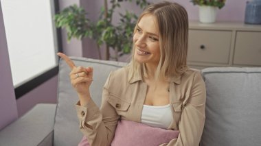Çiçeklerle dolu bir salonda oturmuş gülümseyen ve parmakla işaret eden genç sarışın kadın, olumlu ve misafirperver bir atmosfer sunuyor..