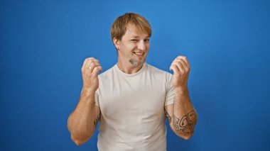 Sakallı ve dövmeli, gülümseyen beyaz bir adam mavi bir duvarın önünde duruyor..