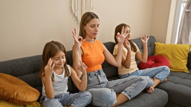 Bir kadın, rahat bir oturma odasında, kanepede iki kızla meditasyon yapıyor..