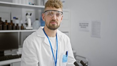 Sakallı ve güvenlik gözlüklü beyaz adam bir laboratuvarda duruyor, profesyonellik ve uzmanlık iletiyor..