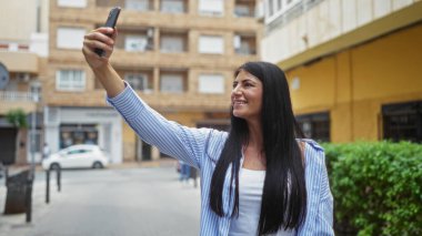 Güzel, genç, İspanyol bir kadın akıllı telefonuyla bir kenar mahallede uzun esmer saçlarını göstererek selfie çekiyor..