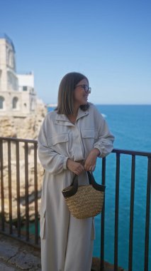 Genç, güzel bir İspanyol kadın, polignano 'da mavi denize bakan güneşli bir günün tadını çıkarıyor. Bir kısrak, puglia, İtalya, günlük giysiler giymiş ve elinde bir kamış torba tutuyor..