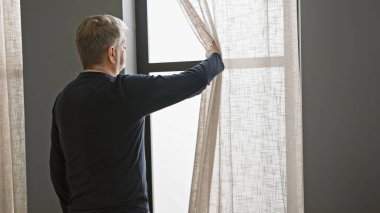 Orta yaşlı, siyah tişörtlü, beyaz saçlı bir adam evde yatak odası penceresinden dışarı bakıyor, yalnızlığı ya da tefekküre dalmayı tasvir ediyor..