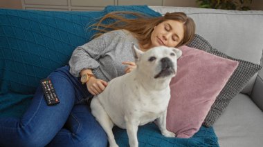 Genç sarışın bir kadın oturma odasında kanepede uyuyor. Yanında beyaz bir köpek oturuyor..