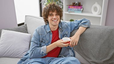 Kıvırcık saçlı, gülümseyen genç bir adam evdeki kanepede, elinde kırmızı bardakla rahatlıyor..