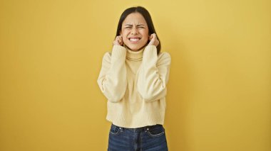 Sarı arka planda kulaklarını kapatan süveterli genç bir İspanyol kadın rahatsız edici bir ifade sergiliyor..