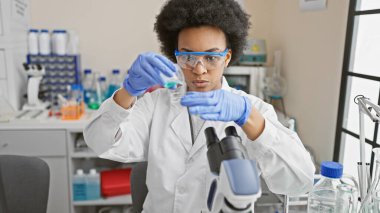 Odaklanmış Afrikalı kadın bilim adamı test tüplerini bir laboratuvar ortamında inceliyor, profesyonelliği ve sağlık alanındaki gelişmeleri resmediyor.