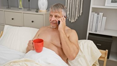 Beyaz çarşaflı bir yatak odasında otururken elinde kırmızı bardakla akıllı bir telefonla konuşan beyaz saçlı bir adam.