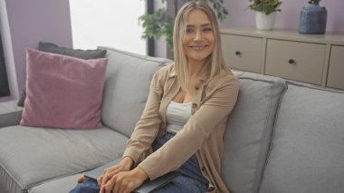 Güzel, genç, sarışın bir kadın rahat bir şekilde gri bir koltukta oturur rahat, rahat, modern bir oturma odasında bitkiler ve yastıklarla, gülümser ve rahat görünür..