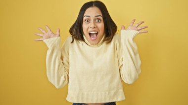 Süveter giymiş, sarı arka planda poz veren, heyecan ve neşe ifade eden İspanyol bir kadın..