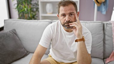 Düşünceli, orta yaşlı, gri saçlı, modern oturma odasındaki bej koltukta oturan İspanyol bir adam..