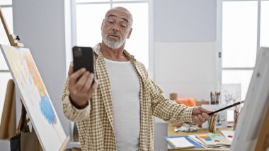 Gri sakallı, tuvale resim yaparken selfie çeken, yaratıcılık ve rahatlık sergileyen olgun bir adam..