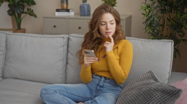 Genç, çekici, sarışın bir kadın evde oturma odasında oturmuş, telefonuyla meşgul olurken düşünceli görünüyor..