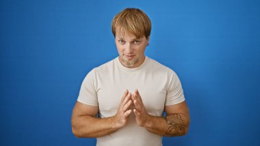 Genç, yakışıklı, sakallı ve dövmeli sarışın bir adam izole edilmiş mavi bir duvara karşı güvenle poz veriyor..