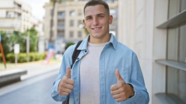 Sırt çantalı neşeli genç bir adam güneşli bir şehir sokağında olumlu ve kendine güvenen bir şekilde başparmak kaldırıyor..